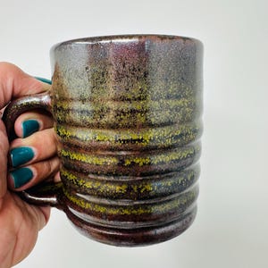 May include: A handmade ceramic mug with a dark brown, green, and yellow glaze. The mug has a cylindrical shape with horizontal ridges and a curved handle. The mug is held by a hand against a white background.