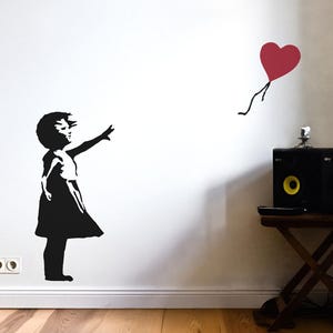 Könnte beinhalten: Schwarz-weiß-Schablone einer jungen Person, die nach einem roten herzförmigen Ballon greift. Das Kunstwerk befindet sich an einer weißen Wand, darunter ist ein Holzboden zu sehen. Im Hintergrund steht ein kleiner Tisch mit Lautsprechern.