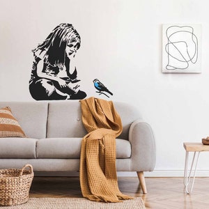 Op de afbeelding: Een grijze bank met een bruin plaid en twee kussens. Een zwart-wit sjabloon van een meisje dat met een blauwe vogel tegen een witte muur zit. Een kleine houten tafel met een wit metalen frame en een kom met noten erop.