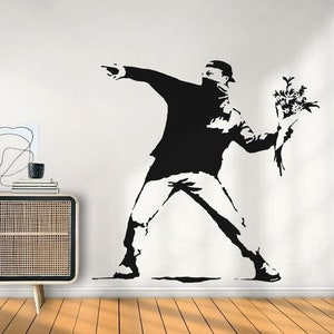 Könnte beinhalten: Schwarz-weiße Silhouette einer Person, die einen Blumenstrauß wirft. Die Person trägt eine Maske und hat den Arm ausgestreckt. Das Bild ist ein beliebtes Street-Art-Werk.