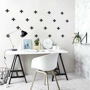 May include: A white desk with a black metal frame and a white chair. The desk has a black lamp, a white clock, and a white vase with a green plant. The wall behind the desk is white with black plus signs.