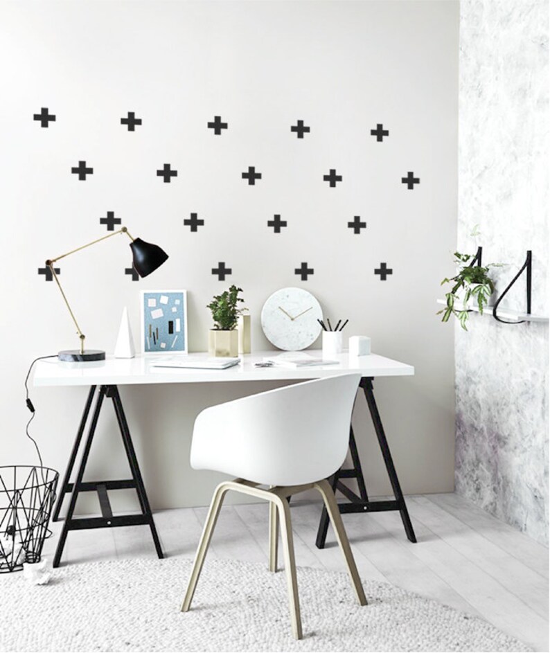 May include: A white desk with a black metal frame and a white chair. The desk has a black lamp, a white clock, and a white vase with a green plant. The wall behind the desk is white with black plus signs.