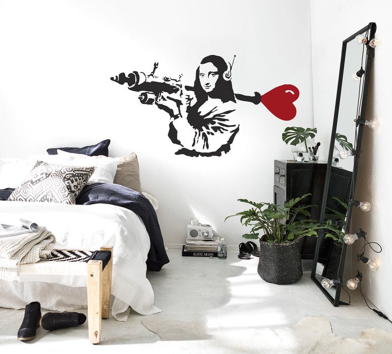 May include: Black and white wall decal of a woman holding a gun with a red heart attached to the barrel. The decal is on a white wall in a bedroom setting with a bed, a plant, and a mirror.