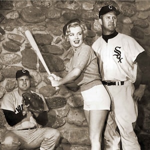 black and white vintage photograph of Marilyn with two uniformed Chicago White Sox players. She is holding a bat poised for a pitch. Behind her is crouched Chicago baseball team catcher and standing next to her is second sox player.