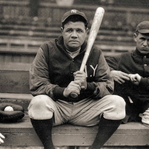 May include: A black and white photo of a baseball player sitting on a bench holding a wooden bat. He is wearing a baseball cap with the letters "NY" on it. The player is wearing a baseball uniform with the letters "NY" on the front.