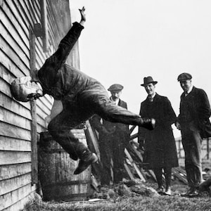 May include: A man in a helmet and goggles is performing a stunt, falling backwards while holding onto a wooden wall. Three men watch the stunt, standing in the background.