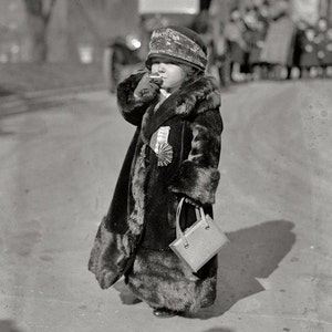May include: A young girl wearing a black fur coat with a matching hat and carrying a small handbag. She is standing on a sidewalk and appears to be looking at something off-camera.