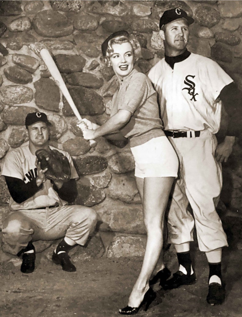 black and white vintage photograph of Marilyn with two uniformed Chicago White Sox players. She is holding a bat poised for a pitch. Behind her is crouched Chicago baseball team catcher and standing next to her is second sox player.