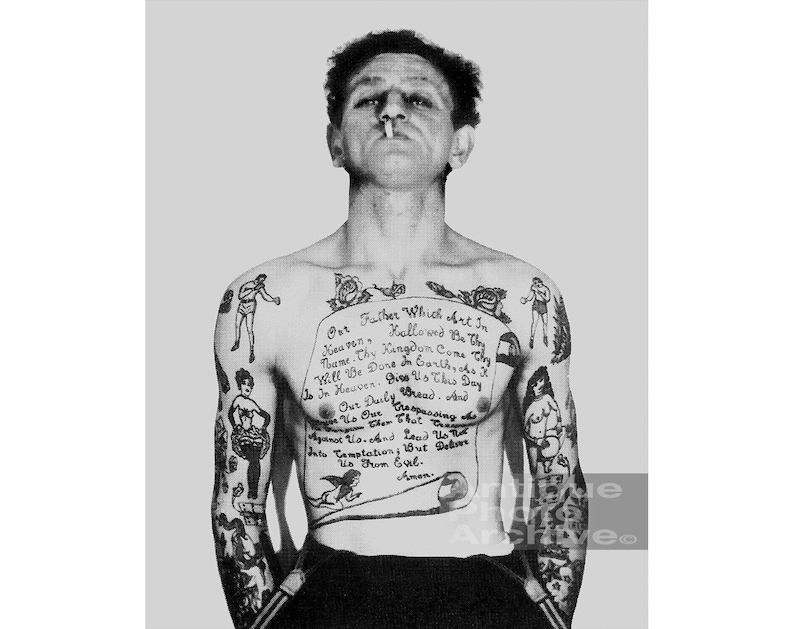 May include: A black and white photo of a man with a full-body tattoo. The tattoo includes a traditional style image of a rose, a boxing scene, and a scroll with the Lord's Prayer written on it.