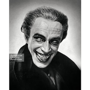 May include: Black and white portrait of a man with a sinister smile. He is wearing a dark suit and has slicked back hair.