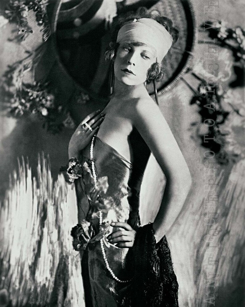 May include: A black and white portrait of a woman wearing a white turban, a beaded necklace, and a velvet dress. She is standing in front of a textured background.