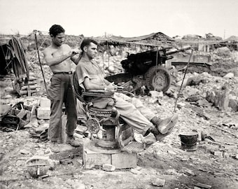 May include: A man sits in a barber chair while another man cuts his hair. The barber chair is in a war-torn area with rubble and debris in the background.