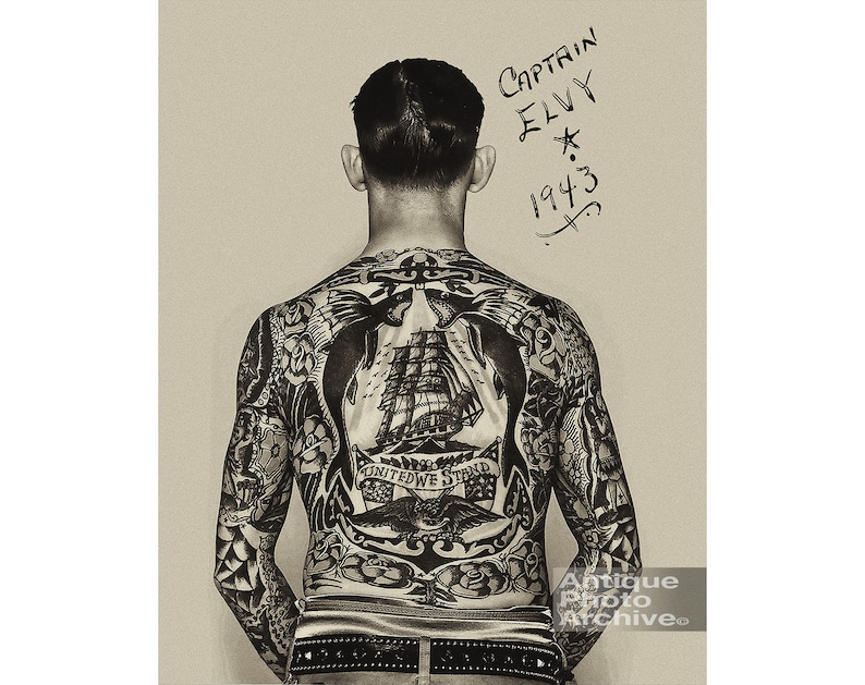May include: Black and white photo of a man's back with a full body tattoo. The tattoo features a ship, an anchor, and the words "United We Stand". The man's name, "Captain Elvy", and the year "1943" are written on the wall behind him.