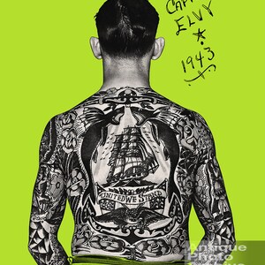 May include: A man with a full back tattoo of a ship, eagles, and an anchor. The tattoo includes the text "United We Stand". The man's back is facing the camera. The text "Captain Elvy 1943" is written in black marker on a green background.