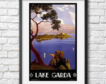 May include: Vintage travel poster advertising Lake Garda in Italy. The poster features a scenic view of the lake with a small island and a castle in the distance. The sky is a light blue with white clouds, and the water is a deep blue. The poster is framed in black and has the text "LAKE GARDA" in large, bold letters at the bottom.