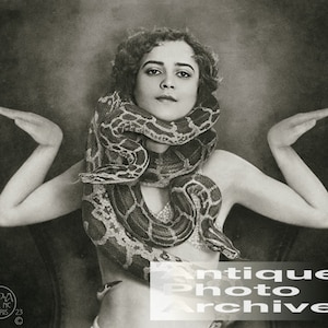 May include: A black and white vintage photograph of a woman with a large snake wrapped around her neck. The woman is wearing a simple outfit and has her arms outstretched. The image is from the Antique Photo Archive.