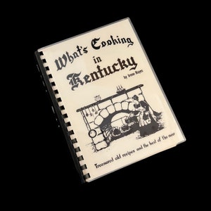 Op de afbeelding: Een vintage kookboek getiteld "What's Cooking in Kentucky" door Irene Hayes. De omslag toont een zwart-witte illustratie van een bakstenen open haard met een vrouw die erin kookt.