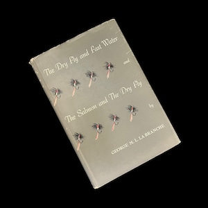 May include: A vintage book titled "The Dry Fly and Fast Water" and "The Salmon and The Dry Fly" by George M. L. La Branche. The cover is gray with illustrations of fishing flies. The book is angled against a black background.