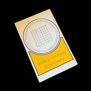 May include: A white book cover with a yellow border and a black and white illustration of the I Ching hexagrams. The title of the book is "BOOK of CHANGES" and the author is "Aleister Crowley".