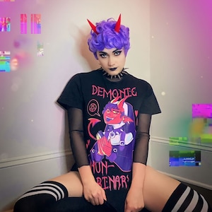 Puede incluir: Una persona con cabello morado y cuernos de diablo rojos lleva una camiseta negra con una imagen de dibujos animados de una monja demonio sosteniendo un libro. La camiseta tiene el texto "Demonic Nun-Binars" impreso en ella. La persona lleva calcetines altos de rayas blancas y negras.