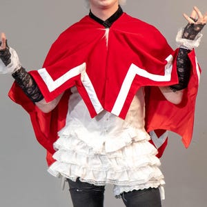 May include: A person wearing a red cape with white accents, a white ruffled skirt, black tights, and black boots. The person is also wearing a white wig with a crown and black gloves. The person is smiling and gesturing with their hands.