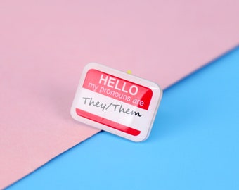 May include: A white name tag with red lettering that says "HELLO my pronouns are They/Them" on a pink and blue background.