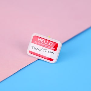 May include: A white name tag with red lettering that says "HELLO my pronouns are They/Them" on a pink and blue background.