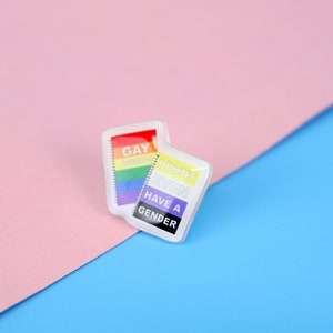 Może przedstawiać: Dwie małe kwadratowe białe szpilki z tęczowymi flagami dumy i tekstem "Gay AF" i "Have a Gender" na różowym i niebieskim tle.