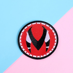 May include: A red and black embroidered patch with a stylized letter "M" in the center. The patch is surrounded by a white border and a black circle.