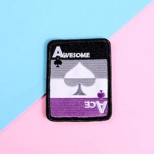 Op de afbeelding: Een zwart-witte patch van een aas van schoppen speelkaart met het woord "AWESOME" boven de aas en het woord "ACE" onder de aas. De patch is op een roze en blauwe achtergrond.