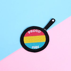 Könnte beinhalten: Eine schwarze Bratpfanne mit einem pansexuellen Pride-Flaggen-Design. Die Pfanne befindet sich auf einem blauen und rosa Hintergrund. Die Pfanne trägt die Aufschrift "Proud Pan".