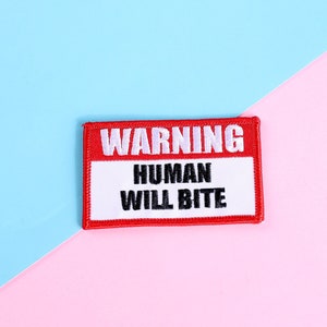 Könnte beinhalten: Ein rechteckiger Aufnäher mit rotem Rand und den Worten "WARNING HUMAN WILL BITE" in schwarzer Schrift auf weißem Grund. Der Aufnäher befindet sich vor einem diagonal geteilten Hintergrund aus Hellblau und Rosa.