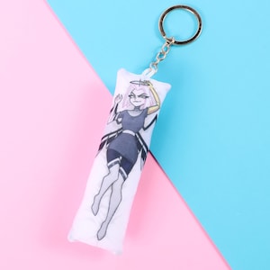 May include: A white keychain featuring a cartoon character on a small pillow-shaped charm. The character is depicted in a dark gray and black outfit with blonde hair. The keychain has a silver ring and chain. The background is split between pink and blue.