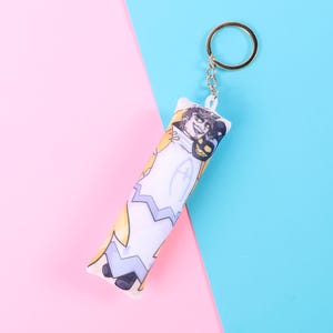 May include: A keychain featuring a cartoon character design. The keychain has a cylindrical shape with a metal ring and chain. The design includes a character in a white, yellow, and purple outfit. The background is a split of pink and blue.