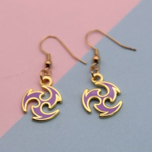 May include: A pair of gold-colored earrings with a purple and gold swirl design. The earrings have a hook closure and are displayed against a pink and blue background.