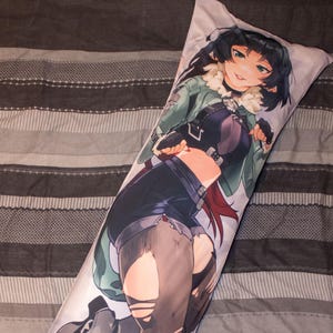 Op de afbeelding: Een lang, rechthoekig lichaamskussen met een kleurrijke anime-stijl illustratie van een vrouwelijk personage. Het personage draagt een groene jas, een zwarte top en een korte broek. Het kussen ligt op een gestreept grijs en wit sprei.