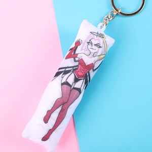 May include: A white rectangular keychain featuring a cartoon illustration of a character in a red corset, black garter belt, and red stockings. The character has pink hair and a halo. The keychain has a gold-colored ring.