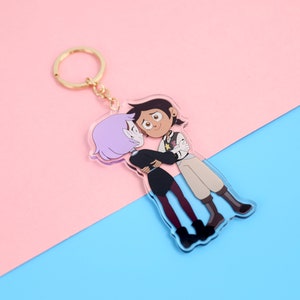 Puede incluir: Un llavero dorado con un dije de acrílico transparente que presenta dos personajes de dibujos animados, uno con cabello morado y el otro con cabello marrón, abrazándose.