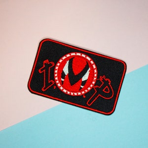 Pode incluir: Etiqueta bordada preta com um círculo vermelho e branco contendo um "V" vermelho estilizado e o texto "TOP" em vermelho.