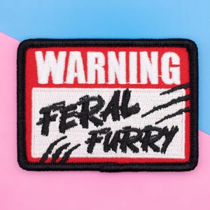 May include: A rectangular embroidered patch with a black border. The top section is red with the white text WARNING. The bottom section is white with the black text FERAL FURRY and black claw mark graphics on the left and right sides.