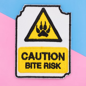 May include: A rectangular embroidered patch with a white background and black border. It features a yellow triangle containing a black animal paw print above a yellow rectangular section with the black embroidered text CAUTION BITE RISK.