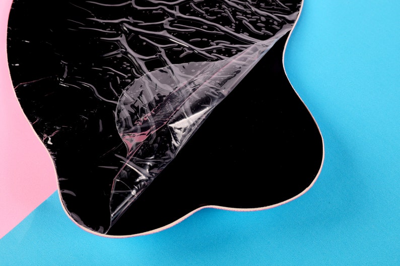 May include: A black, irregularly shaped object with a clear, crinkled protective layer partially peeled back. The background is divided into pink and blue sections, creating a vibrant contrast.