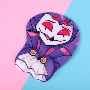 Puede incluir: Alfombrilla de ratón ergonómica morada y rosa con un personaje de dibujos animados con ojos rojos y un cuello blanco con volantes. La alfombrilla tiene un reposamuñecas y está sobre un fondo rosa y azul.