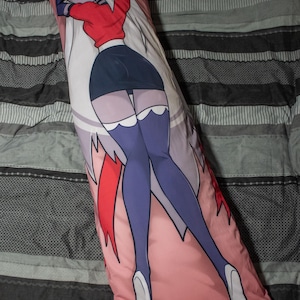 May include: A long, rectangular pillow with a pink background featuring a cartoon character in a red top, black skirt, and purple stockings. The character has white hair and a cross on their forehead.