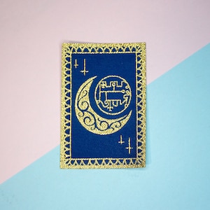 May include: A gold embroidered patch with a crescent moon and a symbol inside the moon on a blue background. The patch has a gold border with a decorative pattern.