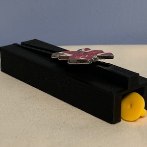 May include: A black plastic 3D printed object with a yellow cap and a silver pin with a red, white, and blue design.