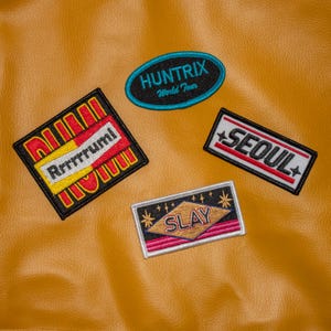 Puede incluir: Un conjunto de parches bordados con diversas formas y diseños. Los parches presentan textos como "HUNTRIX World Tour", "Seoul", "Slay" y "Rrrrrrumi". Los parches son de varios colores, incluyendo negro, rojo, amarillo y blanco, y están sobre un fondo de color mostaza.