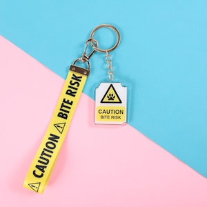 May include: A yellow fabric wrist strap keychain featuring the black text CAUTION BITE RISK and a warning triangle icon, attached to a silver metal ring with a matching rectangular acrylic charm displaying the same warning text and paw print.
