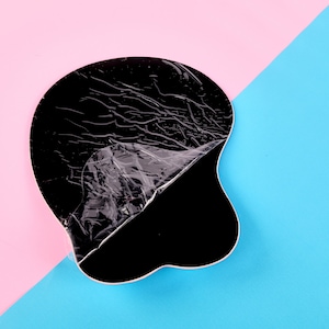 May include: A black, irregularly shaped object with a partially peeled, transparent plastic covering. The object is set against a backdrop of pink and blue diagonal sections, creating a contrasting visual.
