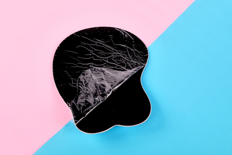 May include: A black, irregularly shaped object with a partially peeled, transparent plastic covering. The object is set against a backdrop of pink and blue diagonal sections, creating a contrasting visual.